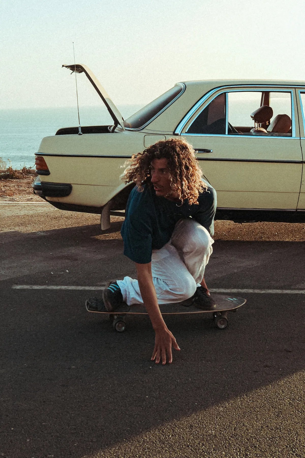 Model von The Sunkissed Rebel skatet in Hockposition vor einem geparkten Oldtimer, trägt das Surf Club T-Shirt in Deep Blue im lässigen Streetstyle.