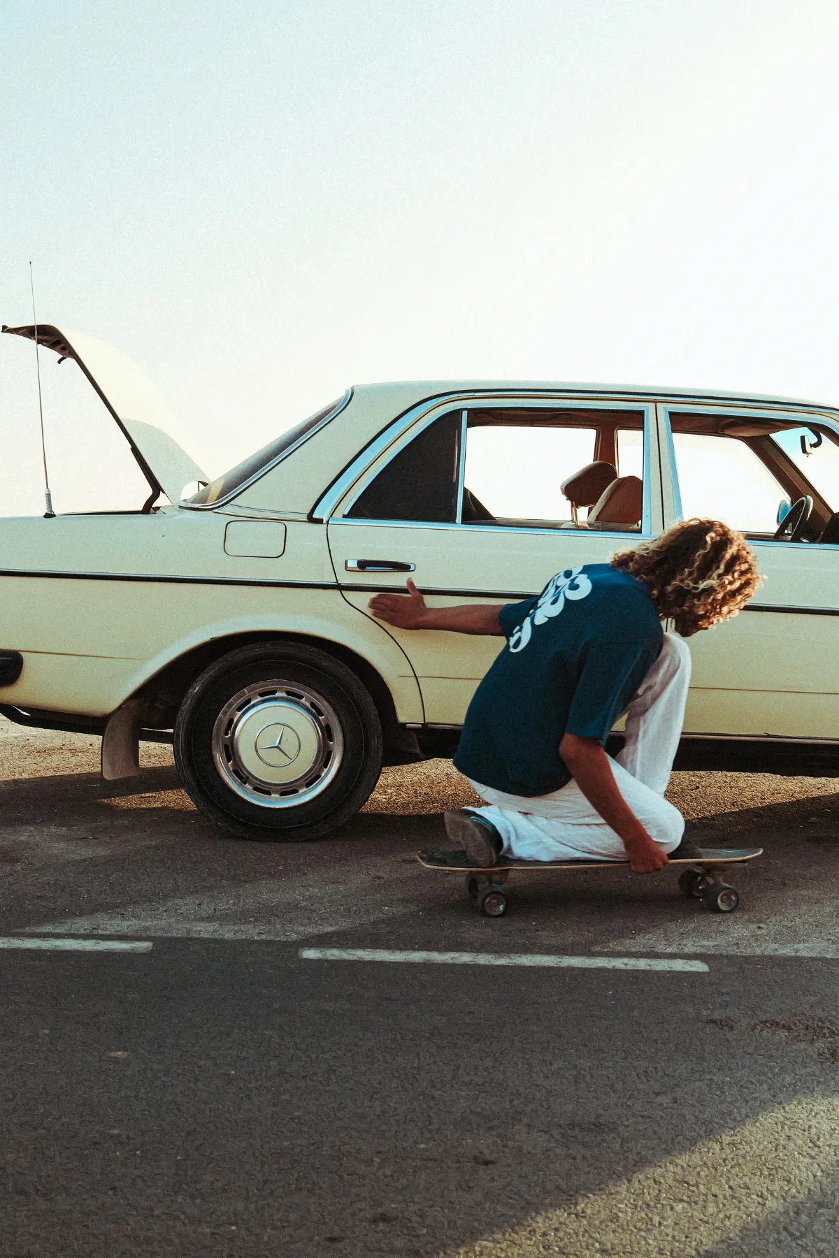 Model von The Sunkissed Rebel rollt tief auf dem Skateboard entlang eines Oldtimers, trägt das Surf Club T-Shirt in Deep Blue mit auffälligem Backprint.