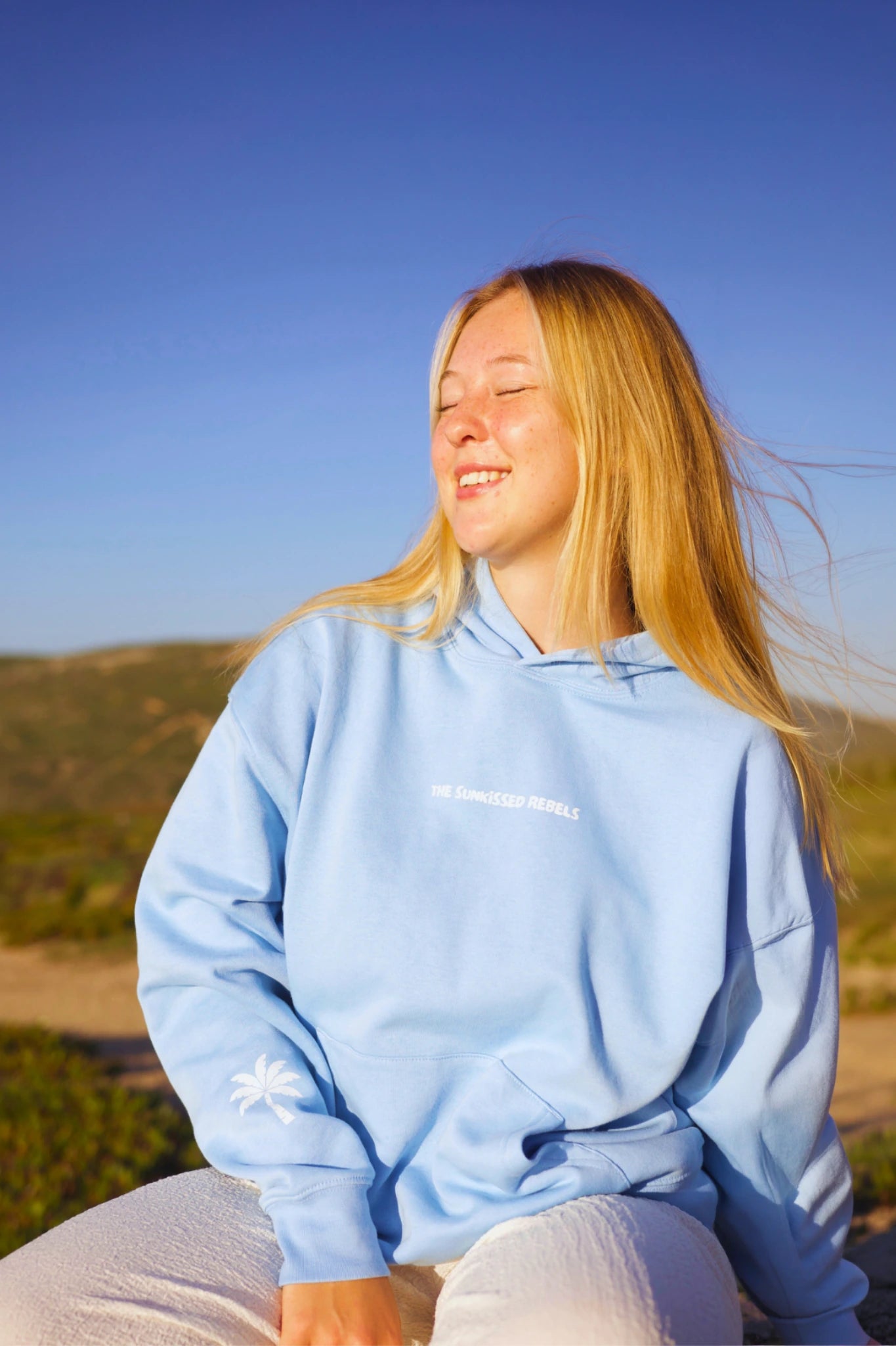 Frau trägt blauen Oversized Hoodie und sitzt entspannt in der Natur bei warmem Licht
