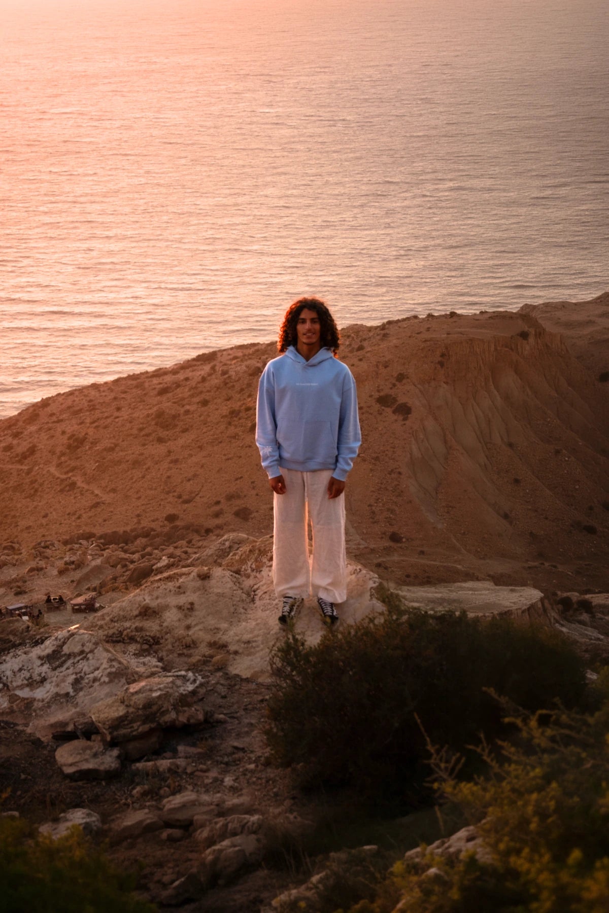 Model im hellblauen The Horizon Hoodie Blue Soul steht auf einer Klippe mit spektakulärem Meerblick bei Sonnenuntergang.
