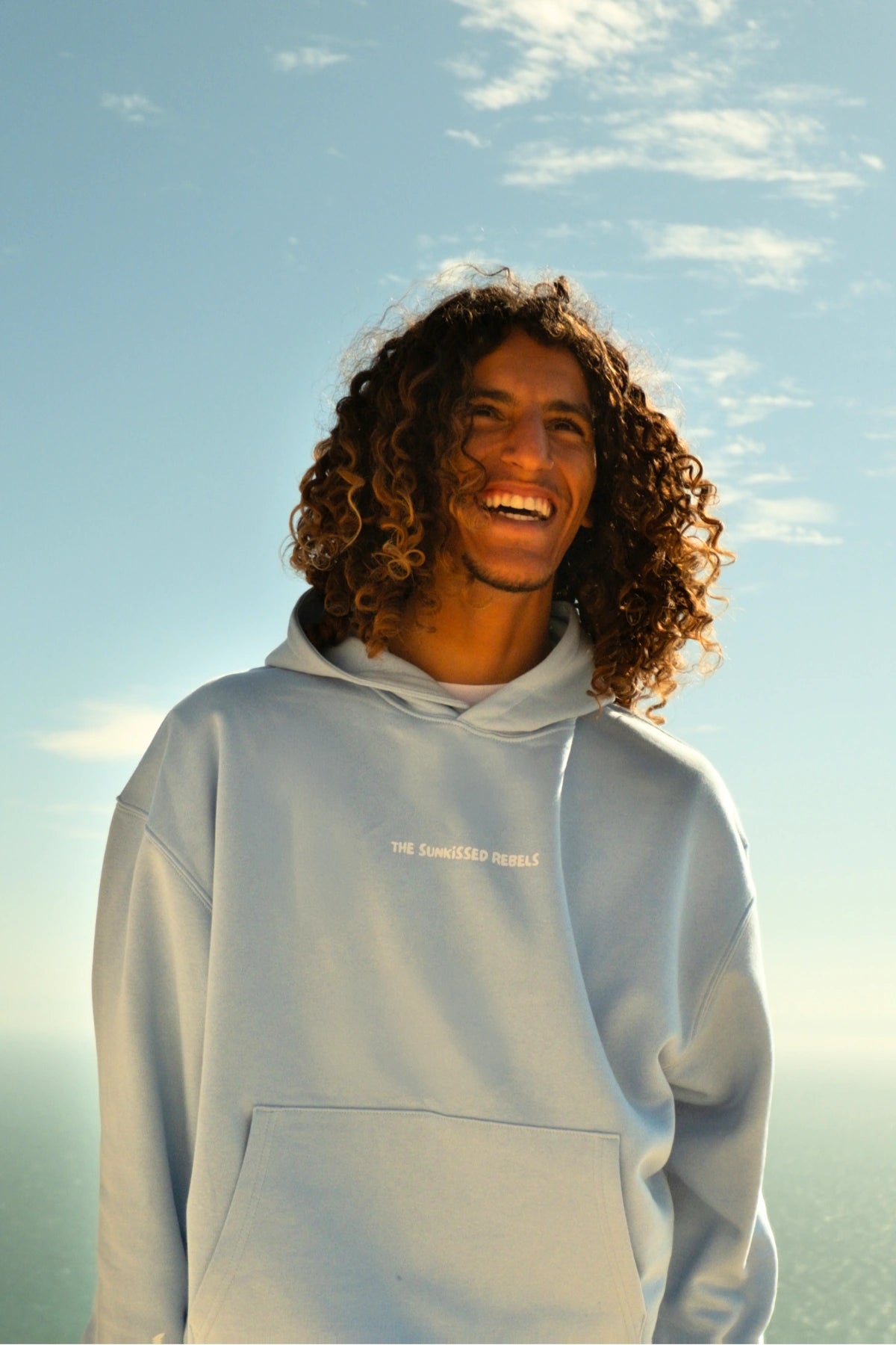 Nahaufnahme eines Models im hellblauen The Horizon Hoodie Blue Soul vor blauem Himmel und sonniger Küstenkulisse.