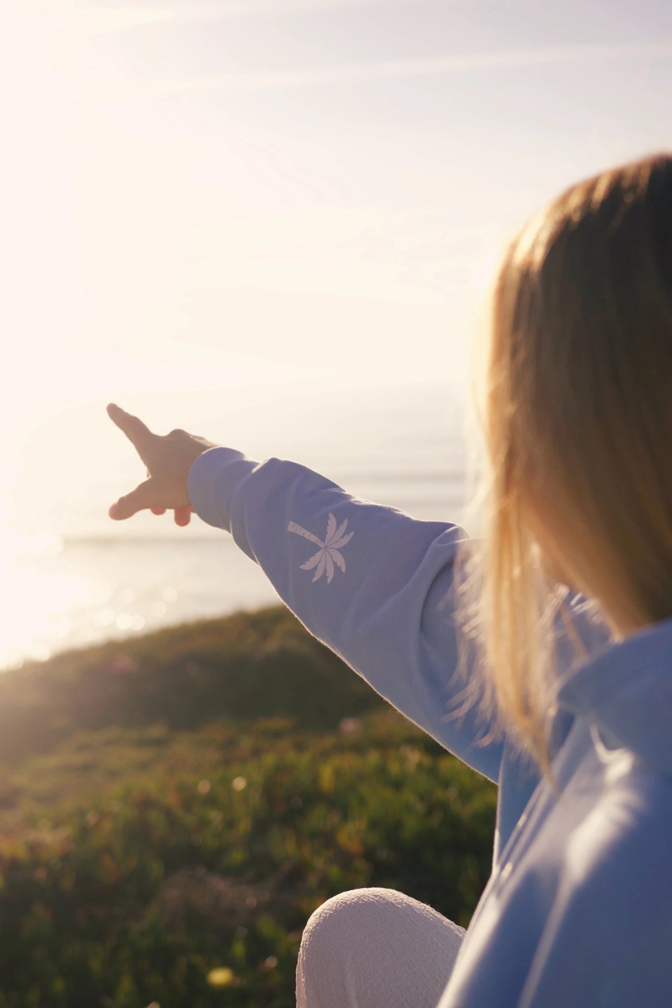 Person zeigt Richtung Meer und trägt blauen Hoodie im Sonnenuntergang mit weichem Licht