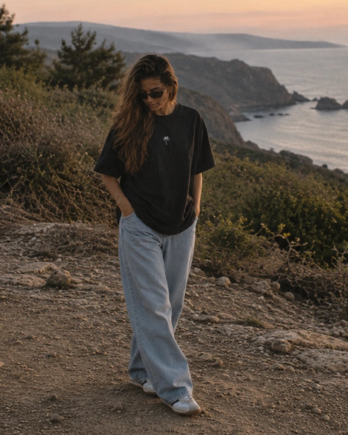 Frau an der Küste im schwarzen Palm Tree Boxy Oversized T-Shirt von The Sunkissed Rebels mit dezenter Palmen-Stickerei und weiter Jeans.