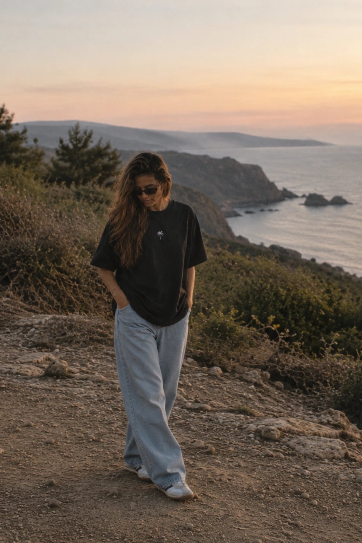 Frau an der Küste im schwarzen Palm Tree Boxy Oversized T-Shirt von The Sunkissed Rebels mit dezenter Palmen-Stickerei und weiter Jeans.