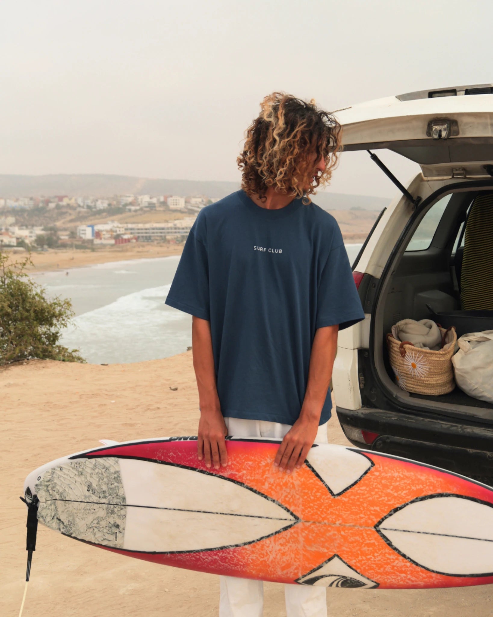 Oversized Surf Club T-Shirt von The Sunkissed Rebels mit Surfboard am Strand fotografiert