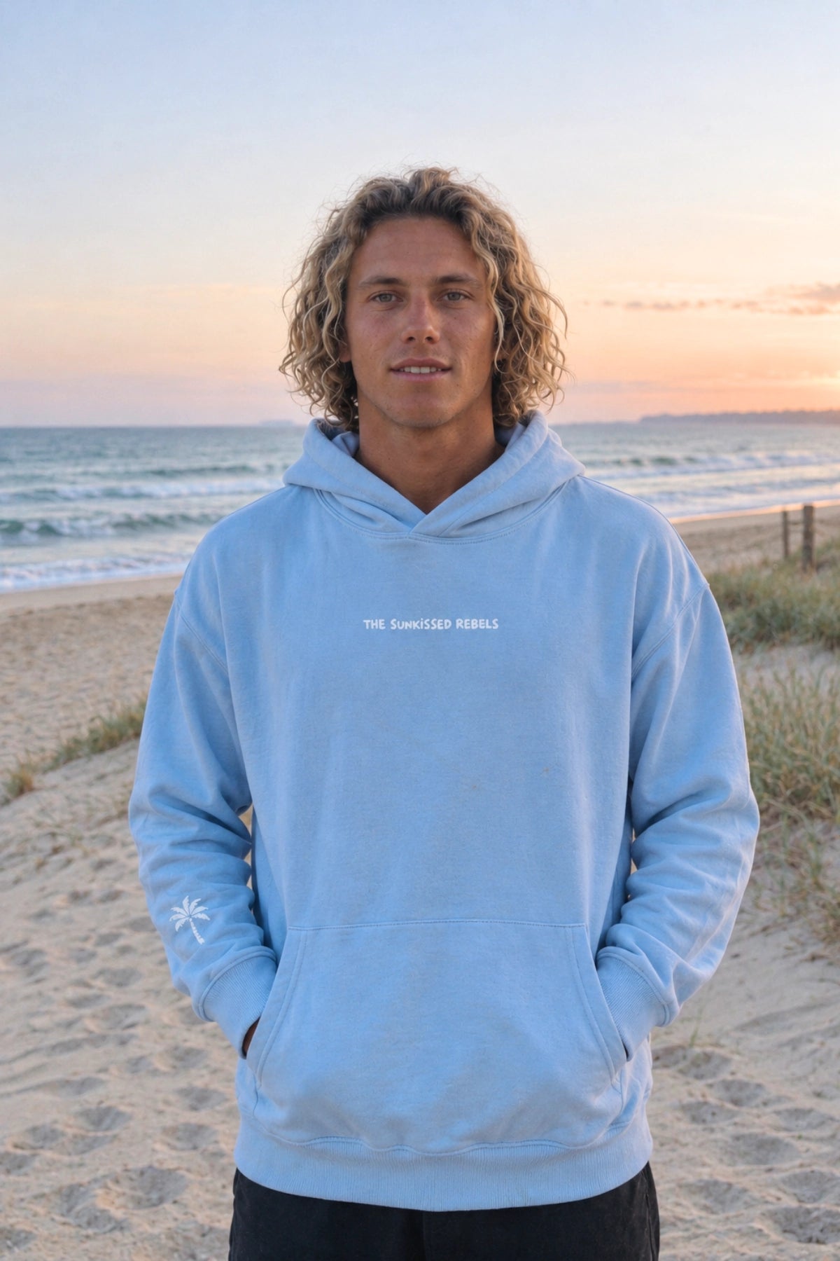 Mann trägt Hellblauer Kapuzenpullover "The Horizon Hoodie - Blue Soul" von The Sunkissed Rebels mit zentralem Logoprint und Palmendetail am Ärmel,