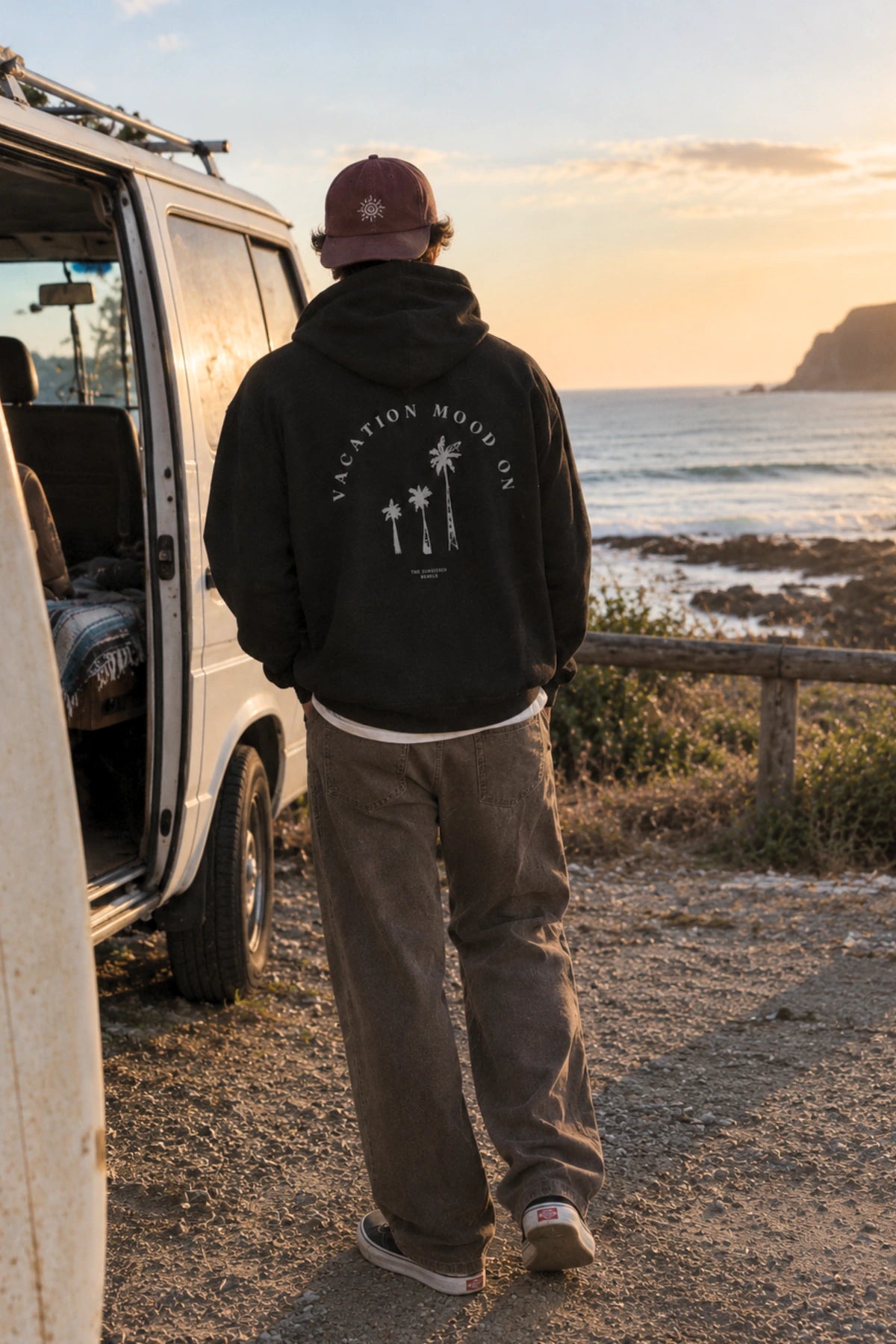Rückansicht des schwarzen Hoodies mit weißem Vacation Mood On Palmen-Print am Campervan mit Blick auf die Küste.