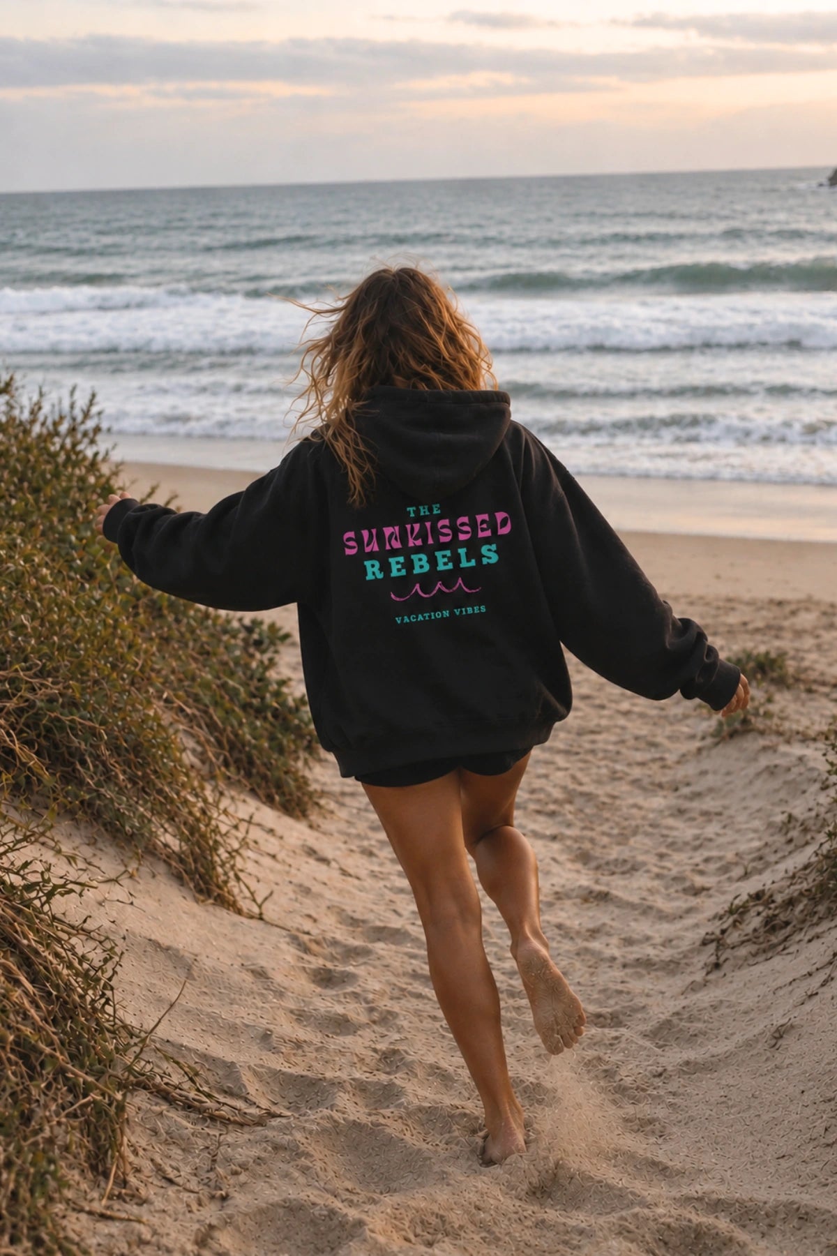 Frau barfuß im schwarzen Vacation Vibes Hoodie am Strand Richtung Meer laufend – The Sunkissed Rebels