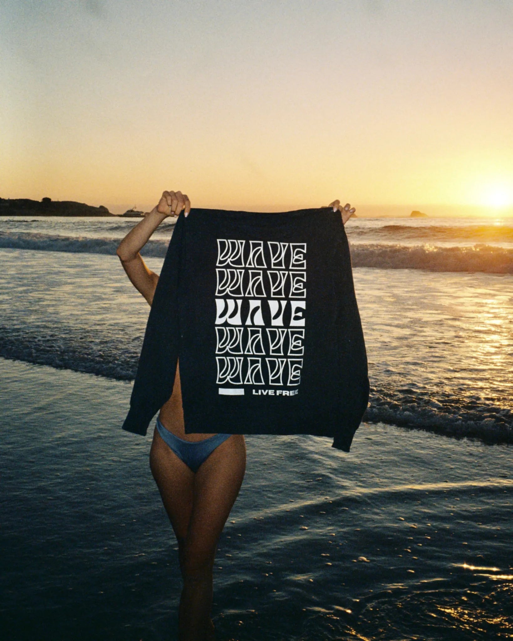 Person hält Wave Live Free Crewneck von The Sunkissed Rebels im Sonnenuntergang am Meer