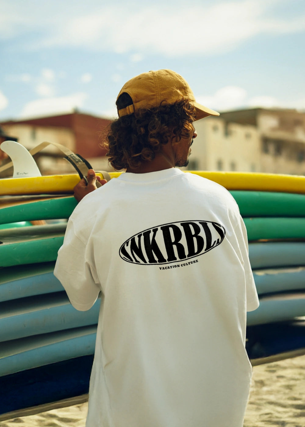 Mann mit Locken und Cap trägt ein weißes T-Shirt mit Backprint und steht vor einer Reihe bunter Surfboards am Strand.