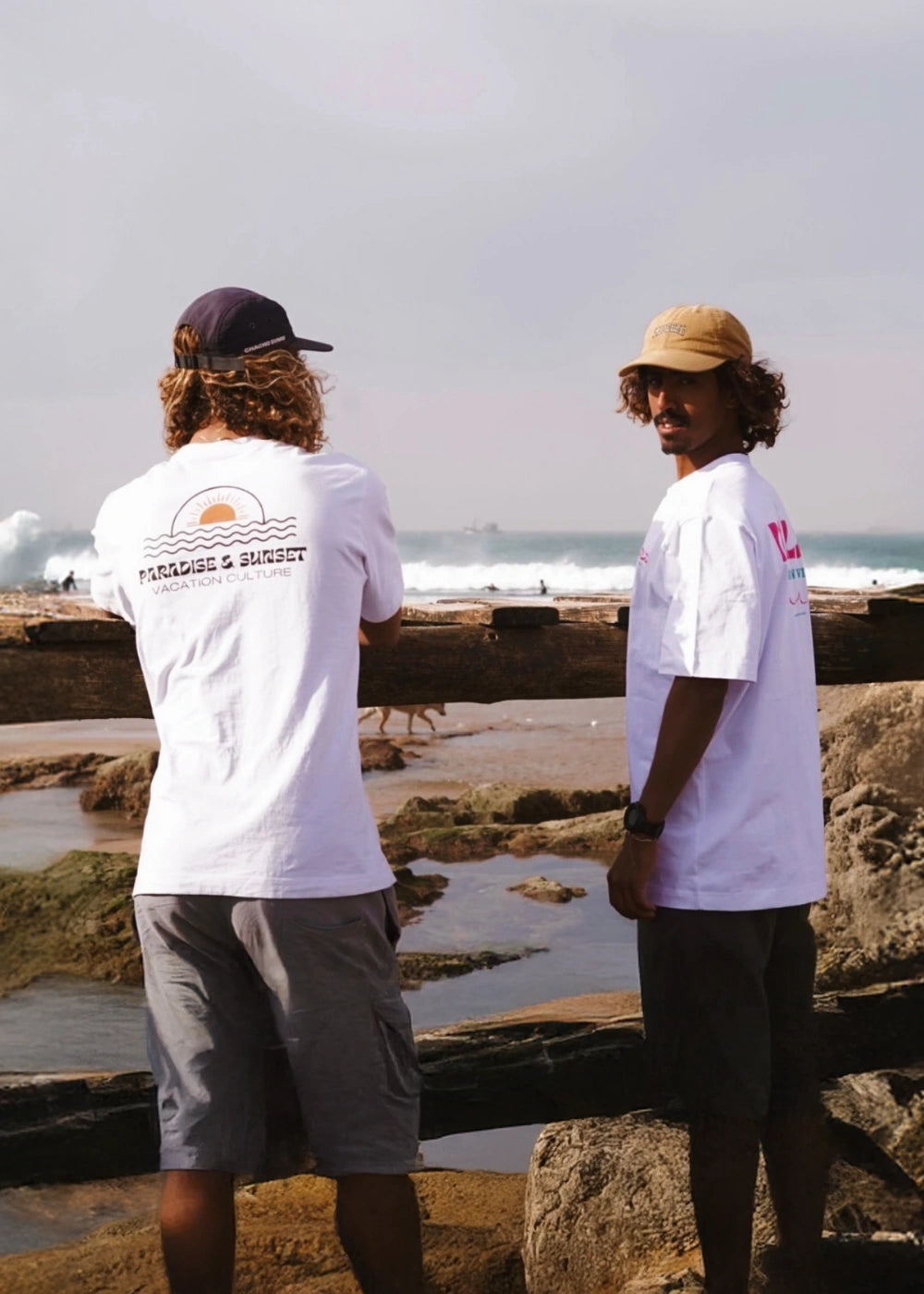 Zwei Männer tragen weiße oversized T-Shirts mit Backprint und stehen auf Felsen mit Blick aufs Meer bei leichtem Wellengang.