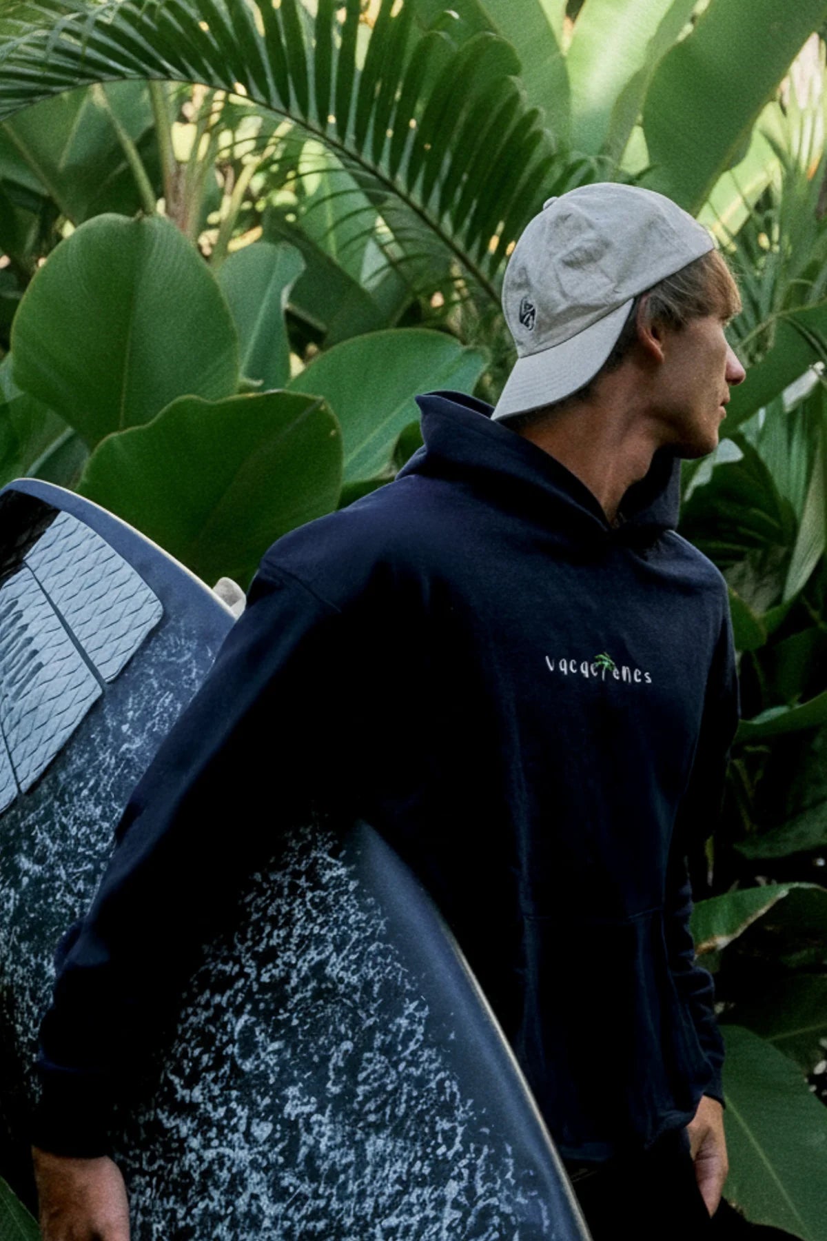 Mann mit Surfboard trägt den Vacaciones Oversized Hoodie Dark Ocean vor tropischen Pflanzen.