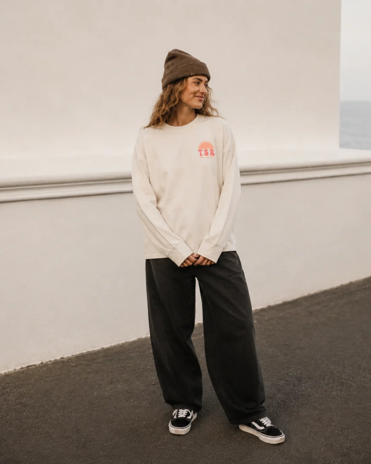 Frau trägt das Vacation Mood Longsleeve in Natural Raw mit TSR-Brustprint, brauner Beanie und weiter schwarzer Jeans.