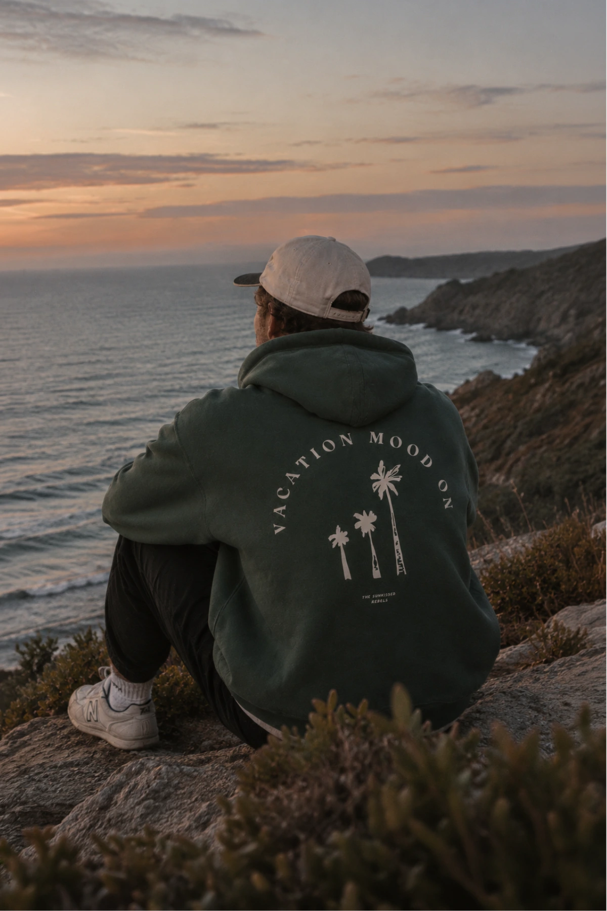 Mann sitzt am Meer im dunkelgrünen Vacation Mood On Hoodie in Green Bay mit weißem Palmen-Rückenprint bei Sonnenuntergang