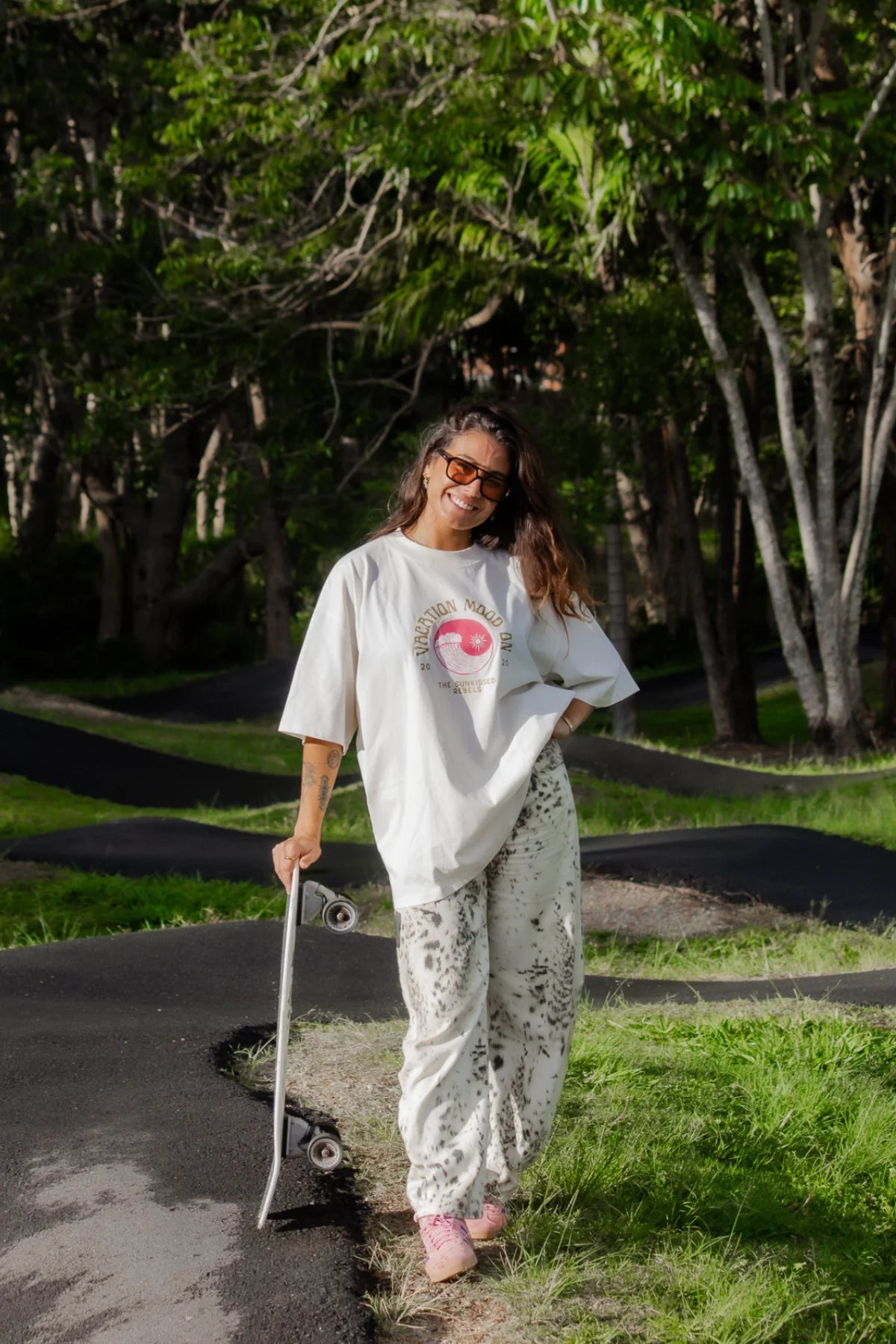 Frau im Oversized T-Shirt Vacation Mood On von The Sunkissed Rebels mit Skateboard im Park