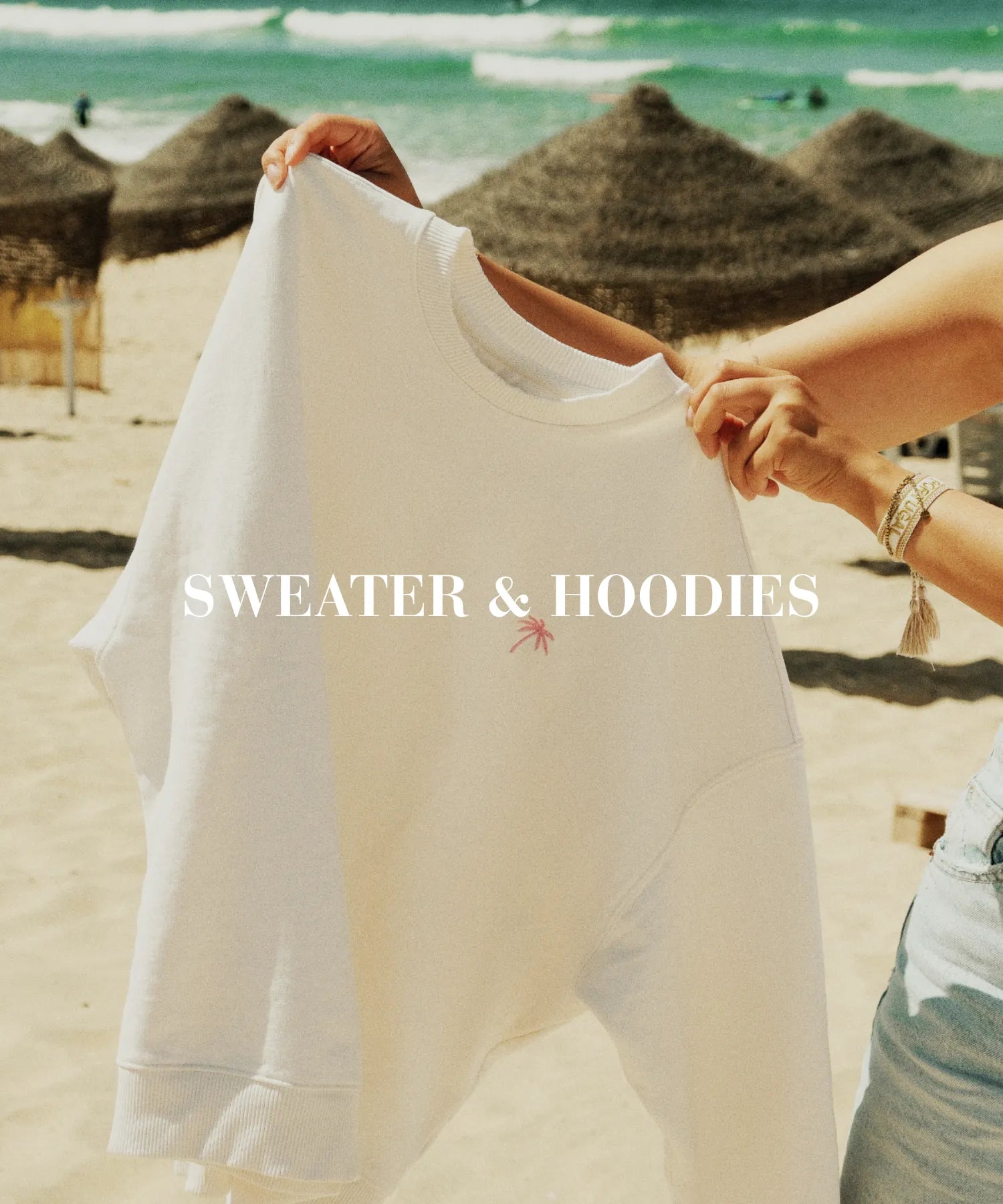 Weißer Sweater mit gestickter Palme wird am sonnigen Strand gehalten – entspannter Sommer-Look mit Stil.