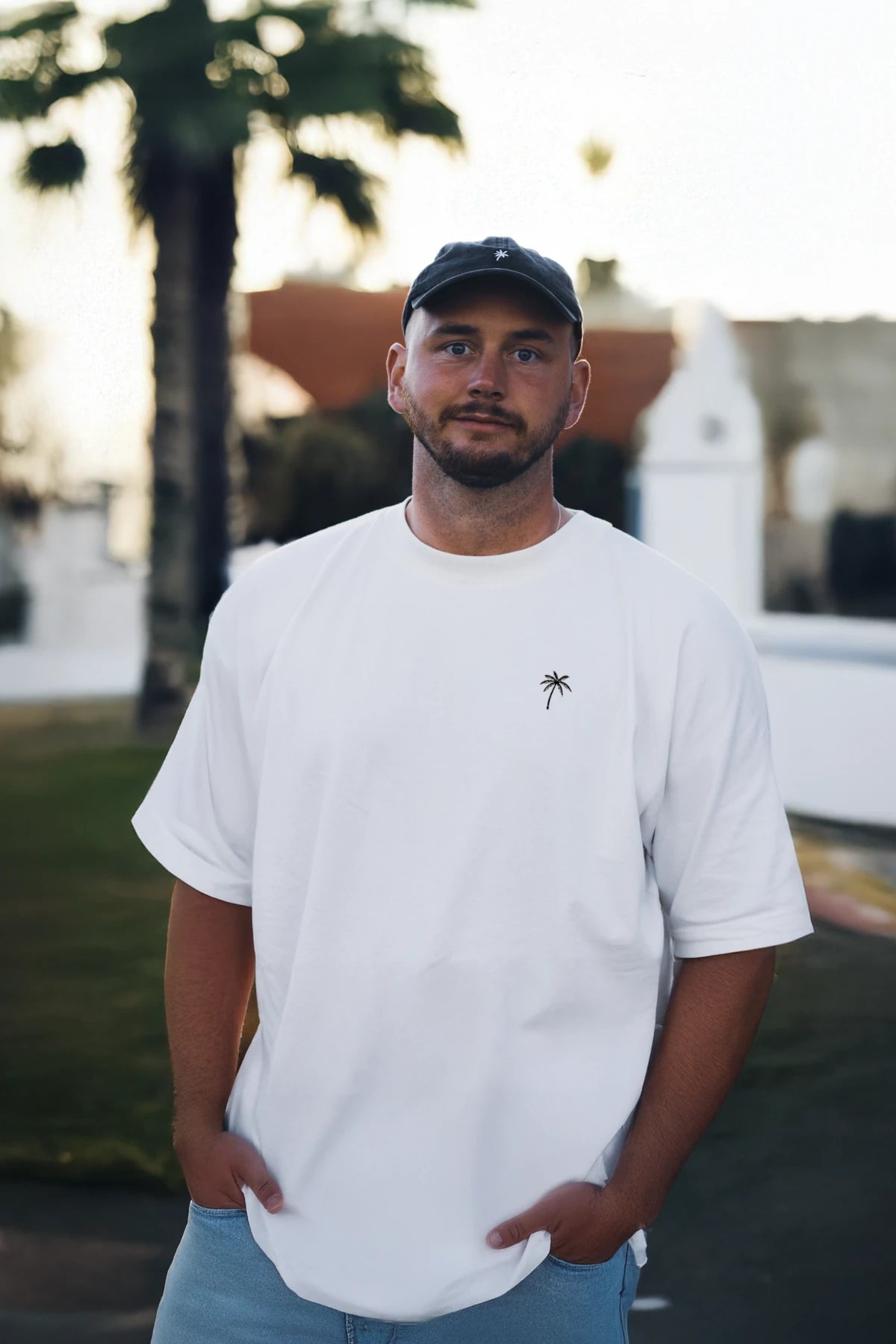 Mann mit weißem Oversized T-Shirt von The Sunkissed Rebels und Palmen-Stick – lässiger Urban Look.